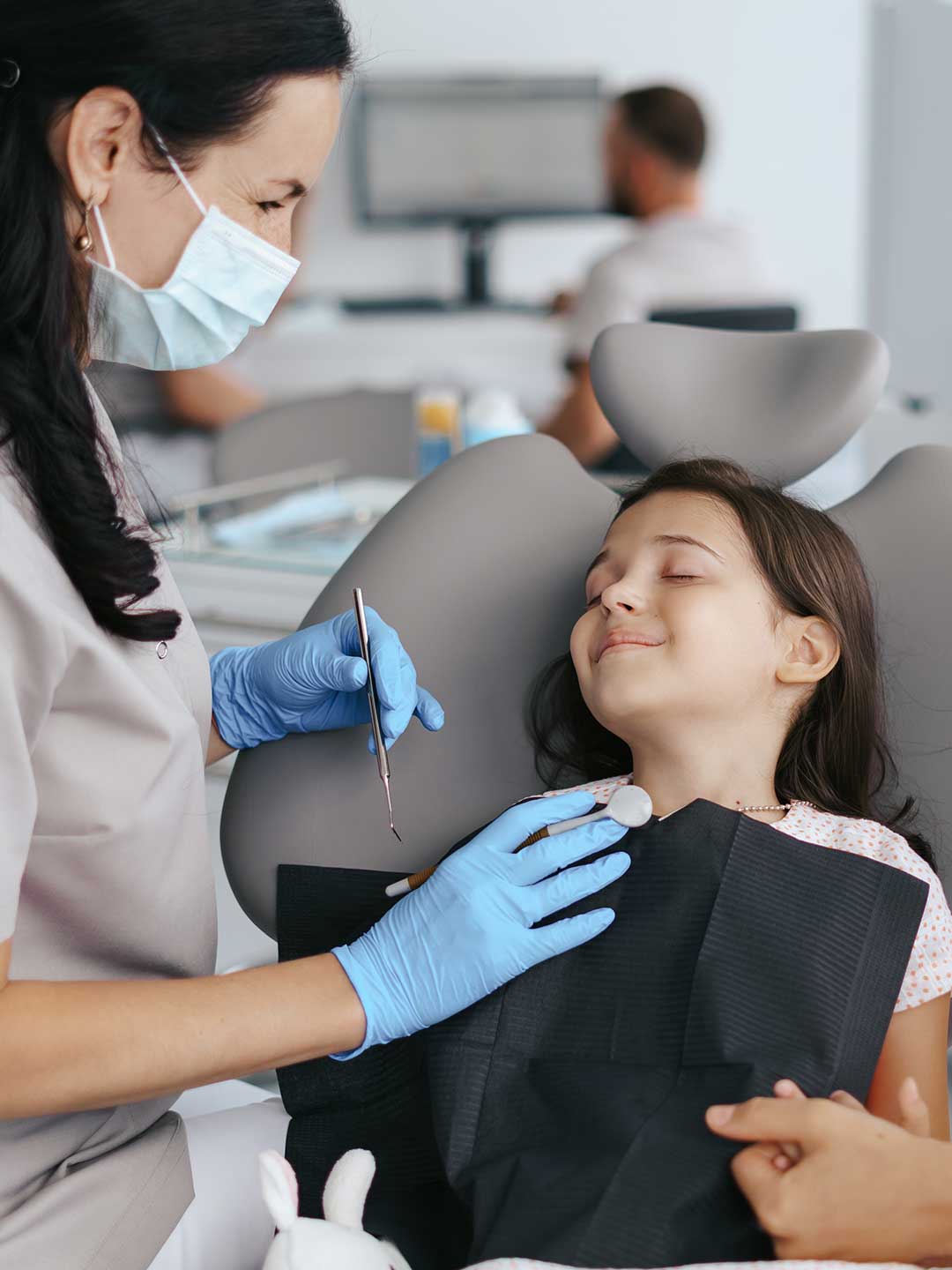 A pediatric dentist gently speaking to a young patient whose eyes are closed, relaxed during treatment.